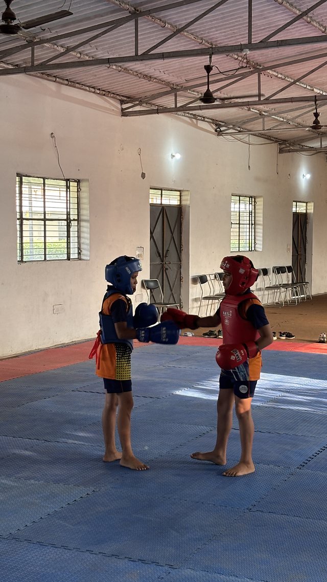 Two students sparring in protective gear on mats in the indoor hall